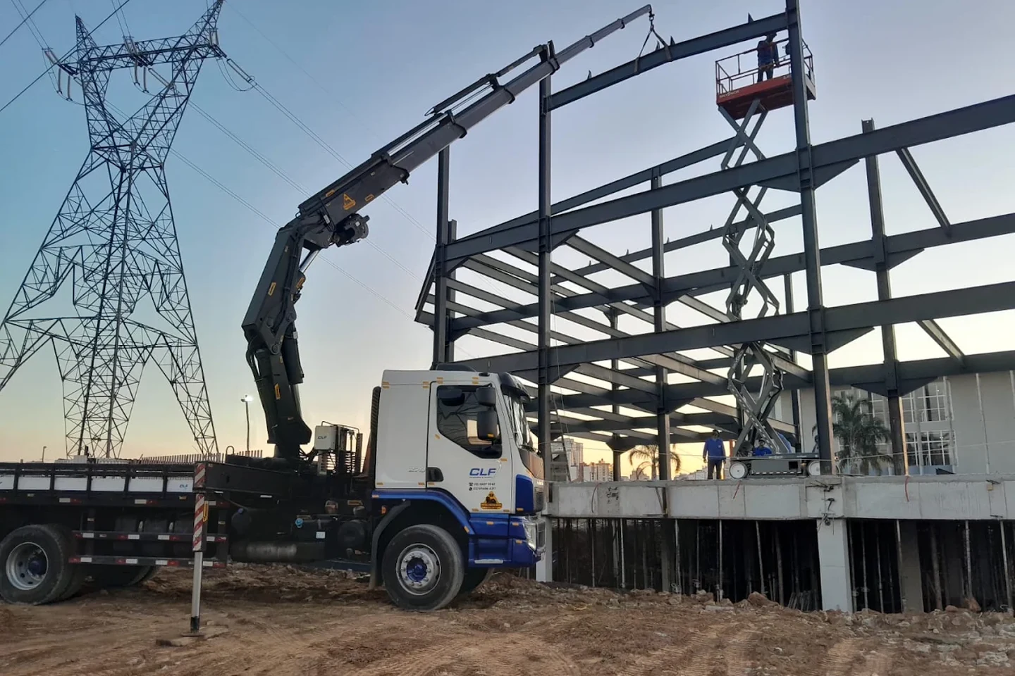 Caminhão munck da CLF com a lança estendida em montagem de estrutura metálica ao entardecer